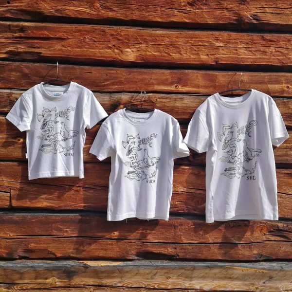 Valkoiset lintukuvioidut lasten t-paidat vanhaa hirsiseinää vasten. Kolme eri kokoista paitaa