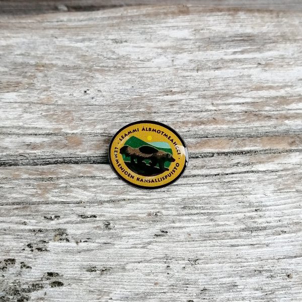 Pieni metallinen pinssi, jossa on Lemmenjoen kansallispuiston logo. Logossa on ahman kuva