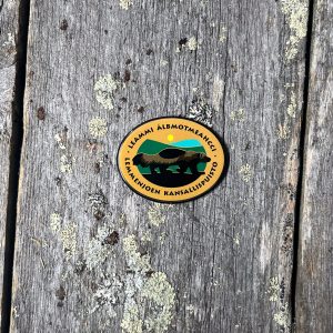 Ovaalinmuotoinen magneetti, jossa Lemmenjoen kansallispuiston logo kuvattuna harmaantuneilla lankuilla
