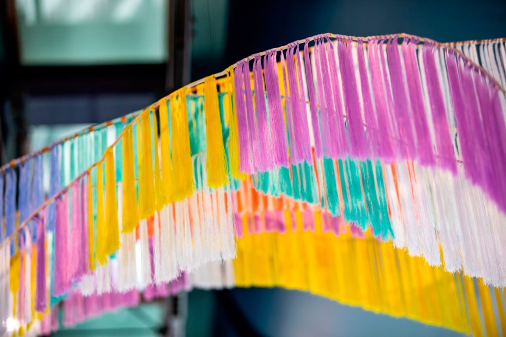 Lähikuva Outi Pieskin teoksesta, jossa roikkuu värikkäitä silkkilankahapsuja vierekkäin ja eri tasoissa.
