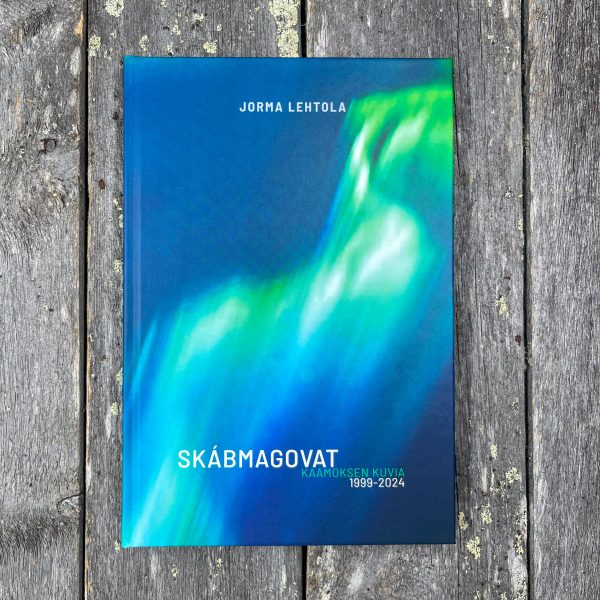 Kuvassa Jorma Lehtolan Skábmagovat, Kaamoksen kuvia 1999-2024 -kirjan kansi kuvattuna harmaantuneella lankulla. Kirjan kannessa vihreät revontulet.