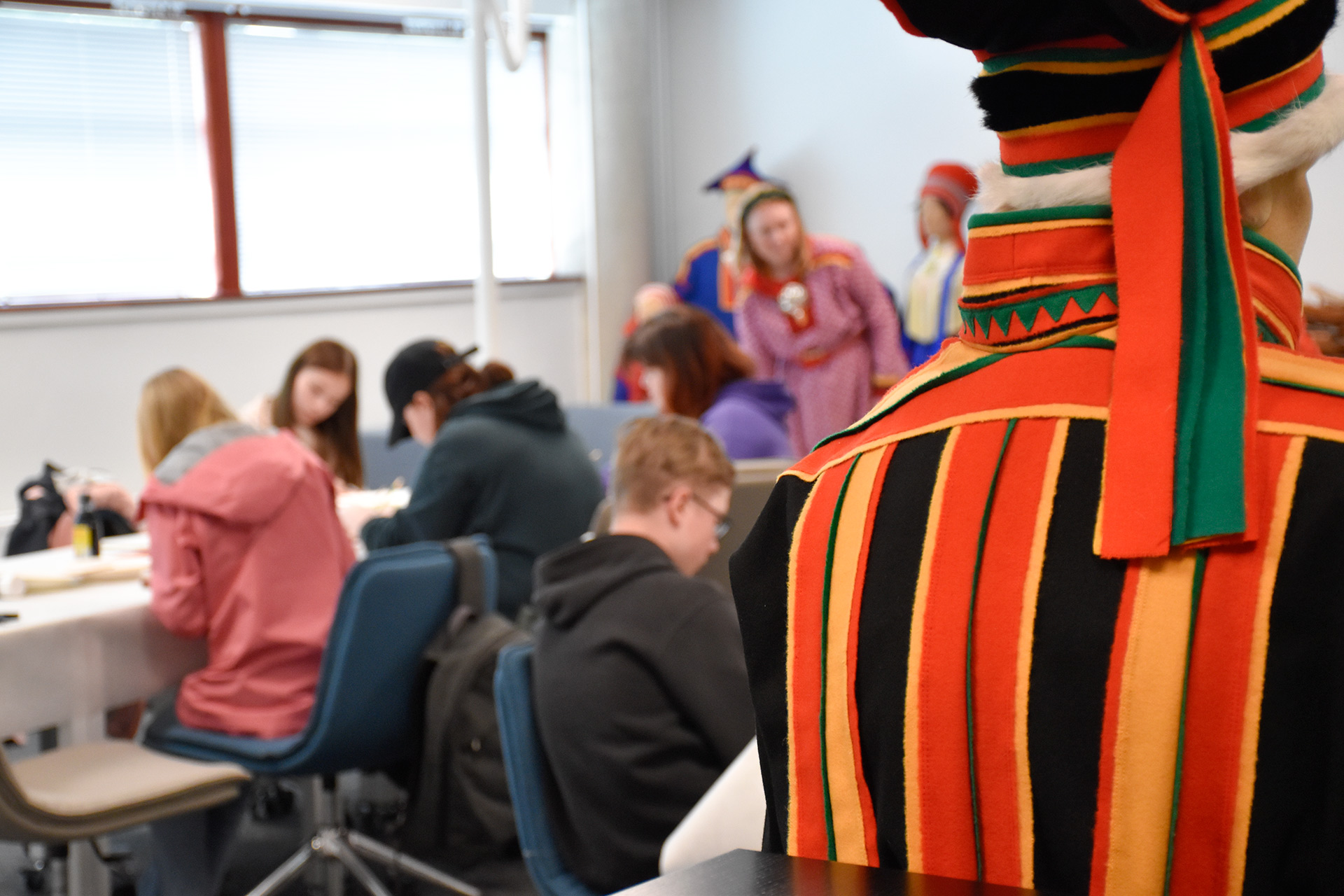 Etualalla inarinsaamelainen miehen puku takaa päin kuvattuna. Taustalla nuoria askartelemassa.