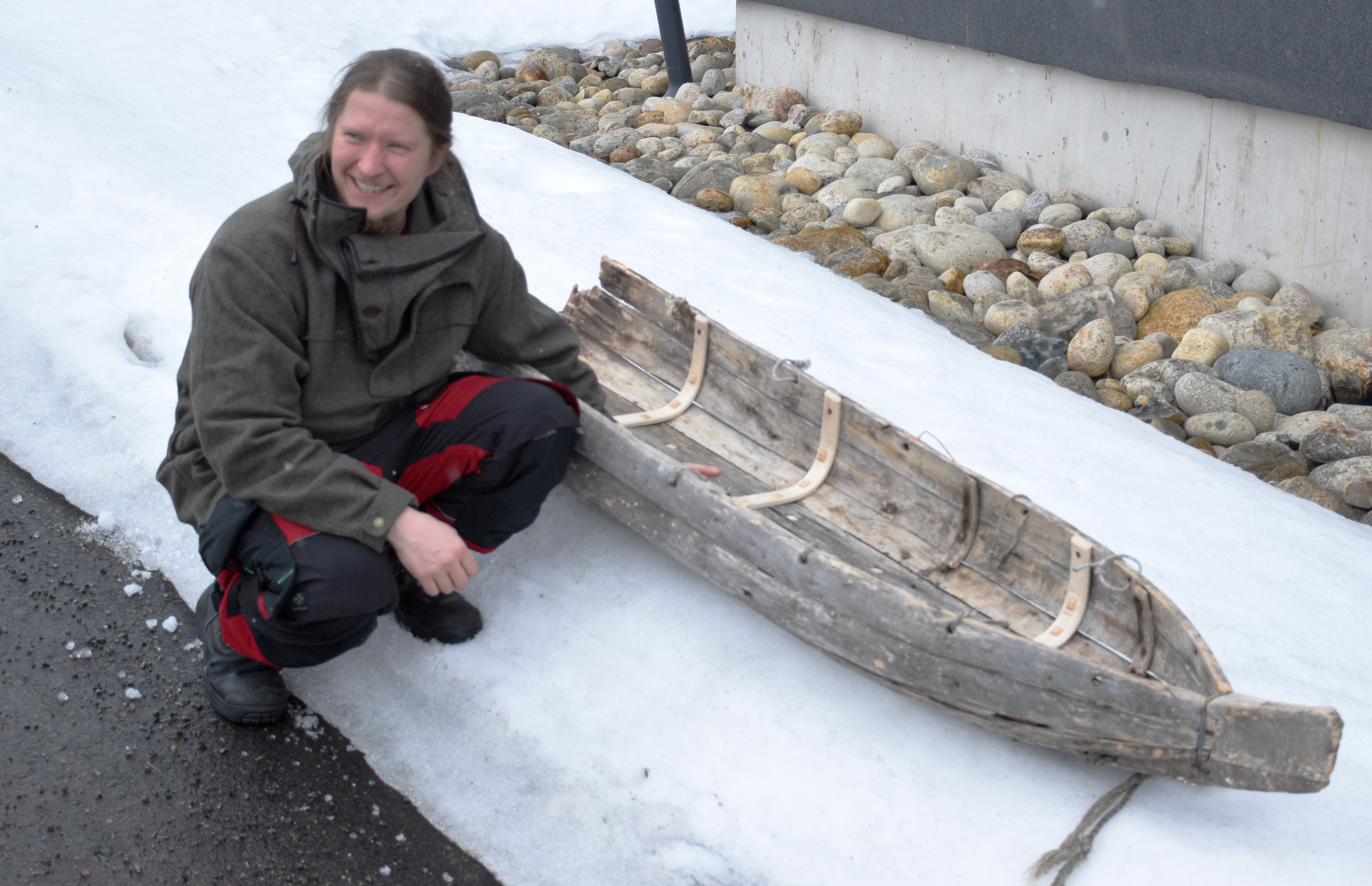 Mies kyykyssä ulkona olevan puusta tehdyn ahkion vieressä