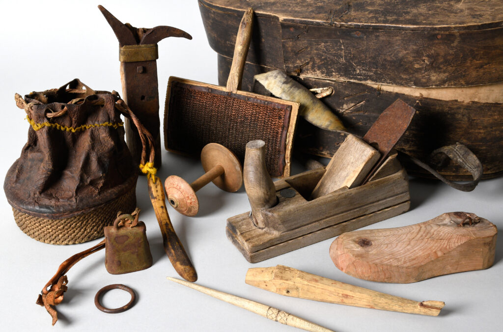 Erilaisia kolttasaamelaisia puisia ja sisnaisia esineitä. Takana kiisa. Etualalla pienempiä esineitä muun muassa höylä, sisnasta ja juuresta tehty pussukka, puinen leikkilentokone